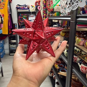 Op de afbeelding: Een helderrode, stervormige ornament met ingewikkelde uitgesneden patronen. De metalen ster wordt in een hand gehouden, wat de driedimensionale vorm benadrukt. De achtergrond suggereert een winkel vol kleurrijke ambachten en decoraties.