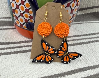 Könnte beinhalten: Ein Paar Ohrhänger mit orangefarbenen, texturierten Ringelblumen-Steckern, die mit schwarz-orangen Monarchfalter-Anhängern mit weißen Punkten verbunden sind und an goldfarbenen Metallhaken auf einer braunen Pappkarte befestigt sind.