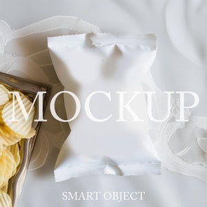 May include: White blank chip bag mockup on a white surface with a silver tray of potato chips in the background. The bag has the text "MOCKUP" and "SMART OBJECT" printed on it.