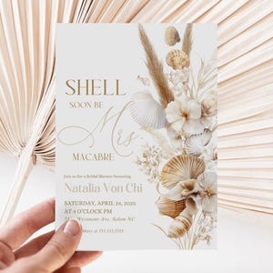 Puede incluir: Una invitación de despedida de soltera blanca y dorada con un diseño de conchas marinas y hierba de la pampa. El texto dice "Shell Soon Be Mrs. Macabre" e incluye la fecha y la hora del evento.