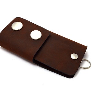 Könnte beinhalten: Ein brauner Leder-Schlüsselhalter mit drei silbernen Druckknöpfen und einem silbernen Schlüsselring.