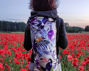 May include: A person wearing a black hoodie with a white and purple tropical print backpack featuring toucans and palm leaves. The person is standing in a field of red poppies.