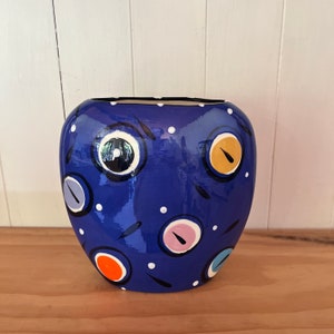 May include: A blue ceramic vase with a unique design featuring colourful circles and white dots. The vase has a rounded shape and a glossy finish.
