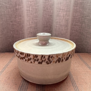 May include: A handmade ceramic lidded bowl. The bowl is off-white with a band of brown speckled design around the rim. The lid and knob are also off-white. The bowl is likely used for food storage or serving.