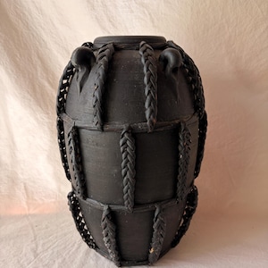 May include: A matte black ceramic vase featuring a rounded body and a narrow opening. The exterior is decorated with a grid of braided black material that wraps vertically and horizontally around the vessel, creating a textured, cage-like appearance.