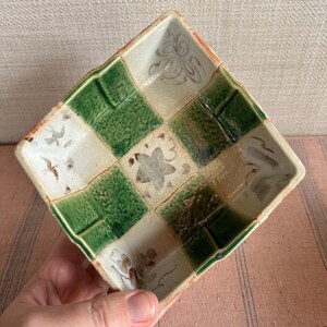 May include: Square ceramic dish with a checkerboard pattern of green and off-white squares. The dish has a raised rim and is decorated with floral and bird motifs. The dish is held by a hand.
