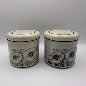 May include: Two ceramic canisters with lids. The canisters are off-white with a floral design in brown. The lids are off-white with a brown ring around the edge. The canisters are cylindrical in shape.