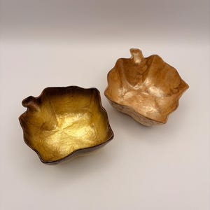 May include: Two leaf-shaped bowls with irregular, textured edges. One bowl features a bright, metallic gold interior finish, while the other bowl displays a lighter, muted bronze or copper-toned interior. Both bowls have a stem-like protrusion.