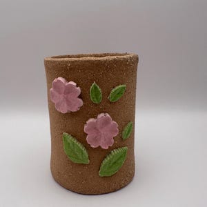 May include: A cylindrical brown ceramic vase decorated with pink flower and green leaf appliqués. The vase has a rough, textured surface and is suitable for holding flowers or other decorative items.