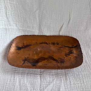 May include: A rectangular copper tray with a textured, uneven edge. The tray has a rich, warm brown colour with darker areas, suggesting a hammered or hand-crafted appearance. The surface has a reflective quality.