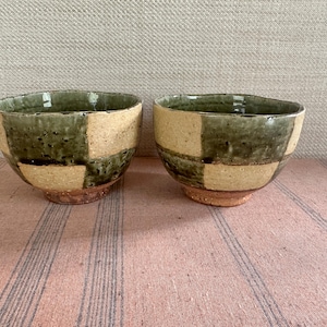 May include: Two handmade ceramic bowls. Each bowl has a distinctive patchwork design of olive green and tan squares. The bowls have a rustic, textured appearance and sit on a pink striped surface.