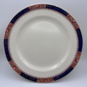 May include: A white ceramic plate with a decorative border. The border features a dark blue band with alternating red and gold floral designs. The plate has a scalloped edge and is suitable for serving food.