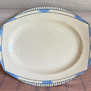 May include: An oval, cream-coloured serving platter with a decorative border. The border features a black and blue geometric design. The platter has a slightly raised edge and is likely made of ceramic or porcelain. Suitable for serving food.