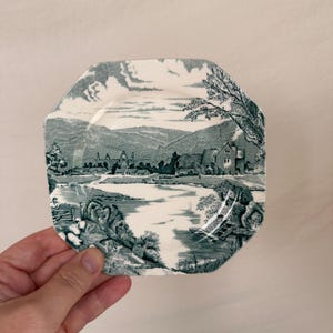 May include: An octagonal ceramic plate featuring a dark green transferware print of a pastoral landscape. The scene depicts a river, a stone building, rolling hills, and trees. The plate has a white background with detailed monochromatic illustrations.