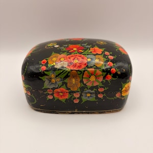 May include: A small, rectangular black box with rounded corners, featuring a hand-painted floral design. The surface is decorated with vibrant red, orange, yellow, and blue flowers, along with green leaves and small berries across the lid and sides.