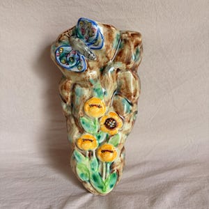May include: A wall-mounted ceramic vase with a textured brown and green finish. The surface features a raised, hand-painted blue and white butterfly at the top and a cluster of four yellow flowers with green stems and leaves at the bottom.