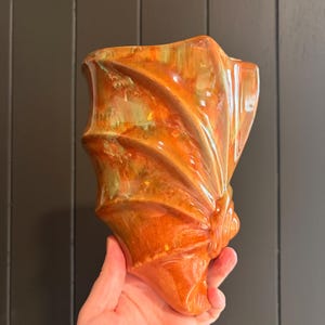 May include: A ceramic wall pocket vase featuring a glossy, multi-toned glaze in shades of vibrant orange, amber, and subtle green. The vessel has a distinct shell-like shape with prominent, curved ridges that fan out from the base to the top.