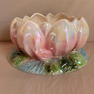 May include: A ceramic bowl shaped like a blooming lotus flower with an iridescent pink and white finish. The base features sculpted green leaves and blue water accents, creating a dimensional appearance with a glossy, reflective surface texture.