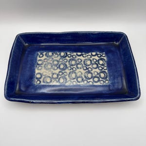 May include: A rectangular ceramic serving tray with a deep blue border and a central design of off-white circles. The tray has a slightly irregular shape, adding to its handmade aesthetic. The glaze has a glossy finish.