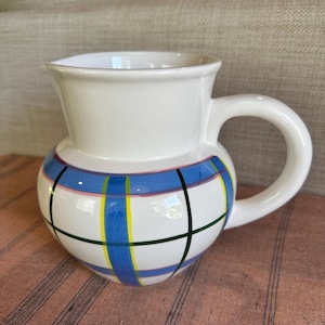 May include: White ceramic jug with a rounded base and a handle. The jug features a blue, yellow, and pink plaid pattern around the middle, with black lines. The top is plain white.