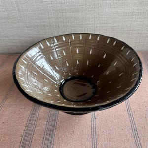 May include: A handmade brown ceramic bowl with a black rim and base. The interior has a pattern of white teardrop shapes. The bowl is likely for serving food or as a decorative item.