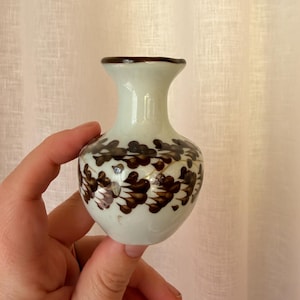 May include: A small, off-white ceramic vase with a narrow neck and a flared rim. The body of the vase is decorated with a repeating pattern of dark brown, leaf-like brushstrokes arranged in horizontal bands around the rounded centre section.