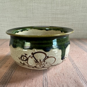 May include: A ceramic bowl with a wide rim and a floral design. The bowl has a cream-coloured base with a crackle glaze and a dark green rim. Brown floral patterns are painted on the exterior.