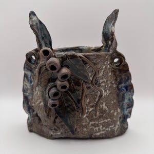 May include: A handmade ceramic vase with a textured, earthy brown exterior. The vase features decorative elements including leaves, berries, and two upward-curving handles. Accents of blue and dark brown add depth to the design, creating a rustic aesthetic.