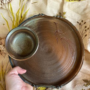 May include: A handmade ceramic serving dish with an attached bowl. The dish is brown with a textured surface and a raised rim. The bowl is a lighter shade of brown with a green interior. A leaf design is etched on the plate.