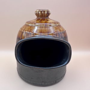May include: A vintage ceramic container with a dark brown, oval opening and a matching lid. The lid and top portion of the container are a lighter brown with decorative detailing. The base of the container is a darker, matte brown.