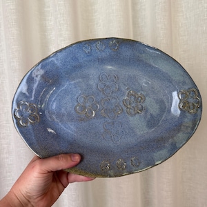 May include: Oval blue ceramic platter with embossed flower patterns. The platter has a textured surface and a raised rim. The flowers are a lighter shade of blue, adding a decorative touch to the serving dish. Measures approximately 30 cm in length.
