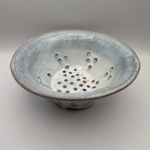 May include: A ceramic colander with a light blue glaze and a speckled interior. The bowl has a wide rim and drainage holes. This handmade colander is designed for washing and draining fruits and vegetables.