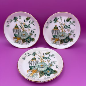 May include: Three white decorative plates with a gold rim. Each plate features a floral design with green and blue-green flowers, leaves, and a decorative urn. The plates are arranged on a purple background.