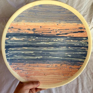 May include: A round ceramic plate with a hand-painted abstract design. The design features horizontal bands of blue, peach, and white brushstrokes. The plate has a cream-coloured rim and a diameter of approximately 25 cm.
