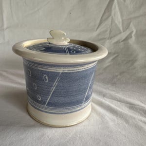 May include: A ceramic jar with a lid, featuring a blue and white design. The jar has a cylindrical shape with a light blue body, decorated with white lines and oval shapes. The lid is blue with white accents and a white, abstract handle.