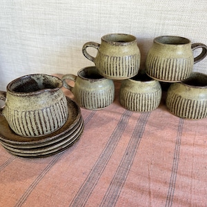 May include: A set of handmade ceramic mugs and saucers. The mugs are light green with vertical brown stripes and small handles. The saucers are stacked. The set is on a pink striped tablecloth.