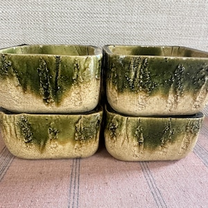 May include: Four square ceramic bowls with a textured, earthy design. The bowls have a light beige base with a green glaze that drips down the sides. The bowls are stacked in two pairs, creating a rustic aesthetic.