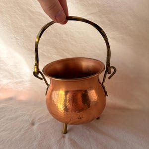 May include: A small, round, hammered copper pot with a wide rim and three short legs. It features a curved, twisted brass handle attached to the sides with metal loops, creating a functional vessel with a metallic, reflective finish.