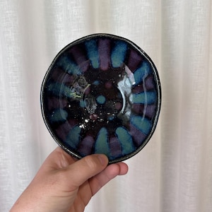 May include: A small, handmade ceramic bowl with a dark blue rim and a vibrant interior. The bowl's interior features a radial pattern of teal and purple hues, creating a unique and artistic design. The bowl is held in a hand.