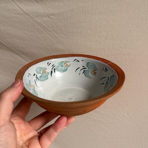 May include: A small, round, terracotta-coloured bowl with a wide, flat rim. The interior is glazed in a creamy white finish and features a hand-painted pattern of light blue, petal-like shapes with small yellow centres arranged around the inner edge.