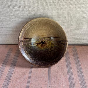 May include: A small, round ceramic bowl with a speckled, earthy design. The bowl features a gradient of colours, transitioning from light tan to deep brown. Dark brown lines and accents add detail to the interior.