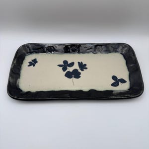 May include: Rectangular ceramic serving tray with a cream-coloured centre and a dark grey border. The tray features dark blue floral designs. The border has a textured, uneven edge. Suitable for serving food or as a decorative piece.