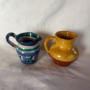May include: Two small ceramic jugs. One is blue with green and white stripes and a handle. The other is yellow and brown with a handle. Both have a spout for pouring.