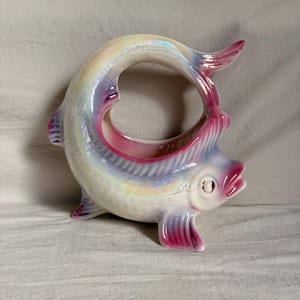 May include: A ceramic fish-shaped planter or bowl with an iridescent, pearlescent finish. The body is curved into a circular shape with a hollow centre. It features painted accents in shades of pink and purple on the fins, tail, and mouth area.