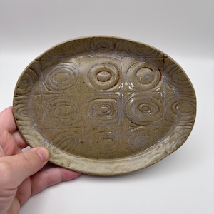 May include: Handmade oval ceramic dish in a speckled tan hue. The dish has a repeating pattern of squares and circles, with a raised edge. The design is carved into the surface, creating a textured appearance. A unique piece for serving or display, measuring approximately 20cm in length.