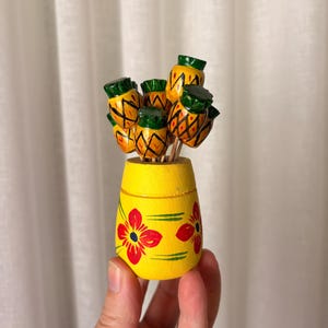 May include: A yellow wooden holder with red and green floral patterns containing several wooden pineapple-shaped cocktail picks. The pineapple picks feature yellow bodies with green tops and brown cross-hatch detailing, standing upright in the container.