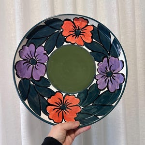 May include: A ceramic plate with a floral design. The plate has a dark green rim and a central olive green circle. The floral pattern includes orange and purple flowers with dark green leaves. The plate is held by a person.