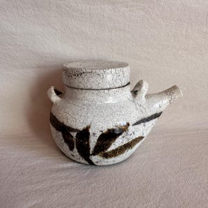 May include: A ceramic teapot with a white crackle glaze finish featuring dark brown leaf-like brushstroke patterns. The vessel has a rounded body, a short spout, two small side handles, and a matching circular lid with a crackled texture.