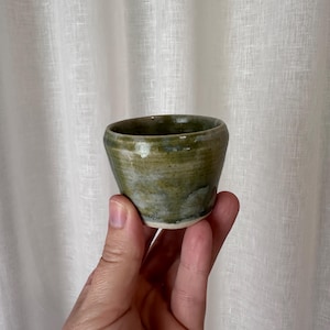 May include: A small, handmade ceramic cup with a rounded shape. The cup has a mottled green and white glaze, with a slightly rough texture. The cup is held in a hand against a white background.