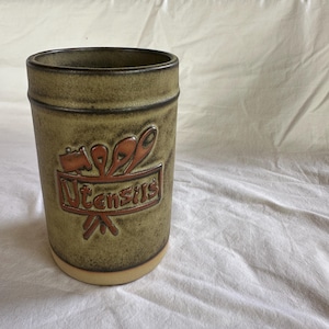 May include: A cylindrical ceramic utensil holder in olive green and brown tones. The holder has a raised design with the word "Utensils" and utensil illustrations in reddish-orange.